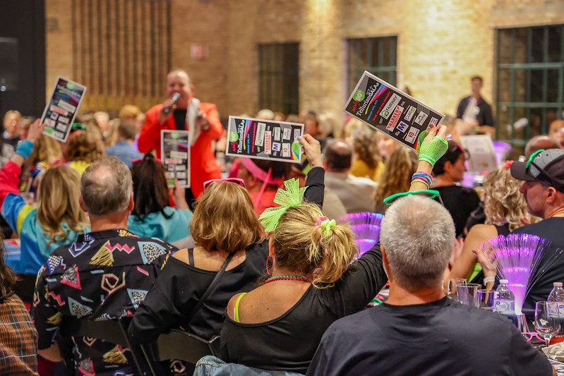 A lively fundraiser crowd holds up bid cards while auctioneer Geoffrey Sandler, in a red jacket, calls the auction at the Tellurian fundraiser