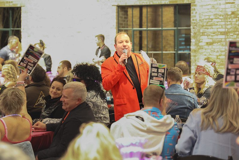 Fundraising auctioneer Geoffrey Sandler in a red jacket on the microphone among guests holding bid cards at the Tellurian fundraiser