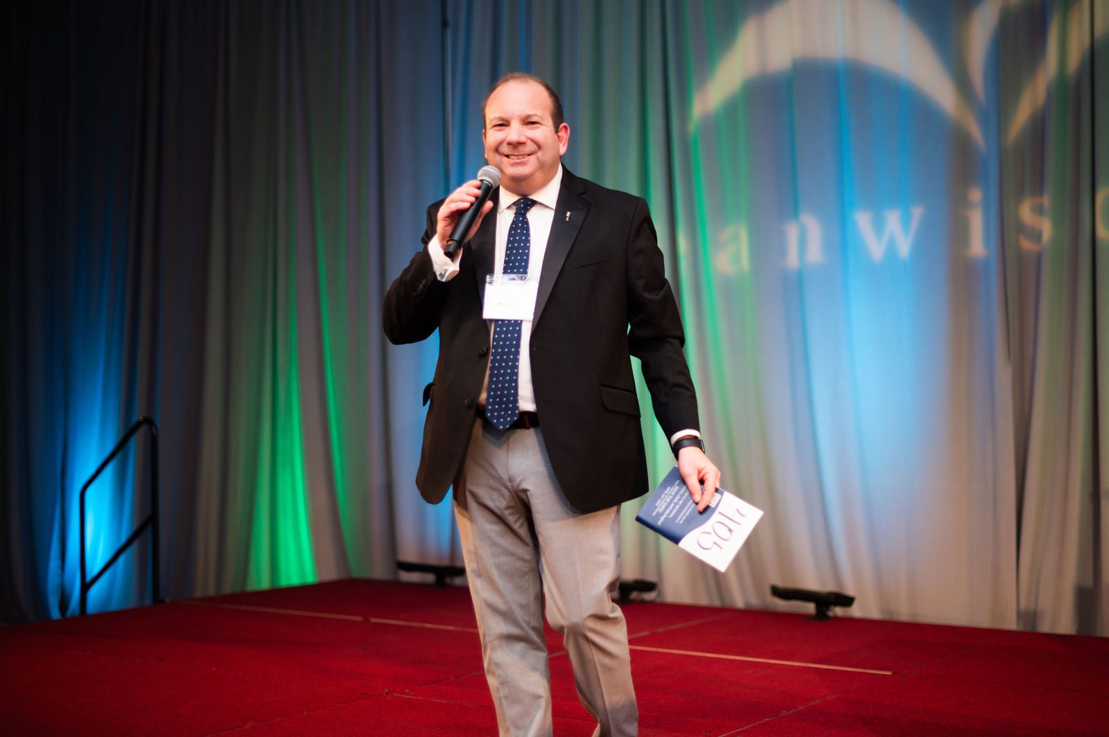 Gala emcee Geoffrey Sandler smiling on stage with a microphone and bid card at Clean Wisconsin's Epicurean Evening