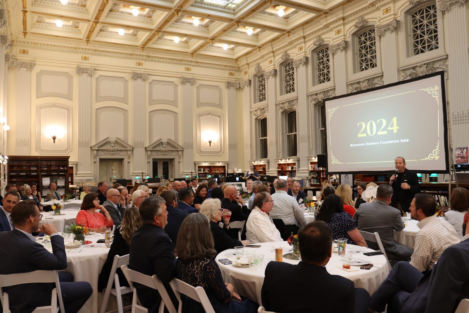 Gala emcee and benefit auctioneer Geoffrey Sandler on the microphone in front of a packed historic hall at the 2024 Wisconsin Bankers Foundation Gala
